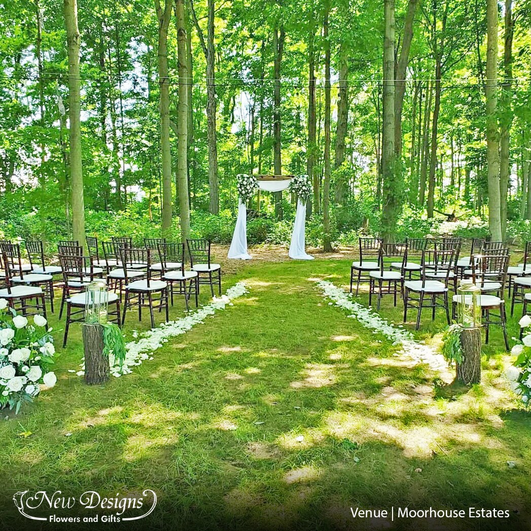 a wooden arch in a forested area with white material, and a white floral installment in each corner, at the end of an aisleway of white padded folding chairs lined with flowers.